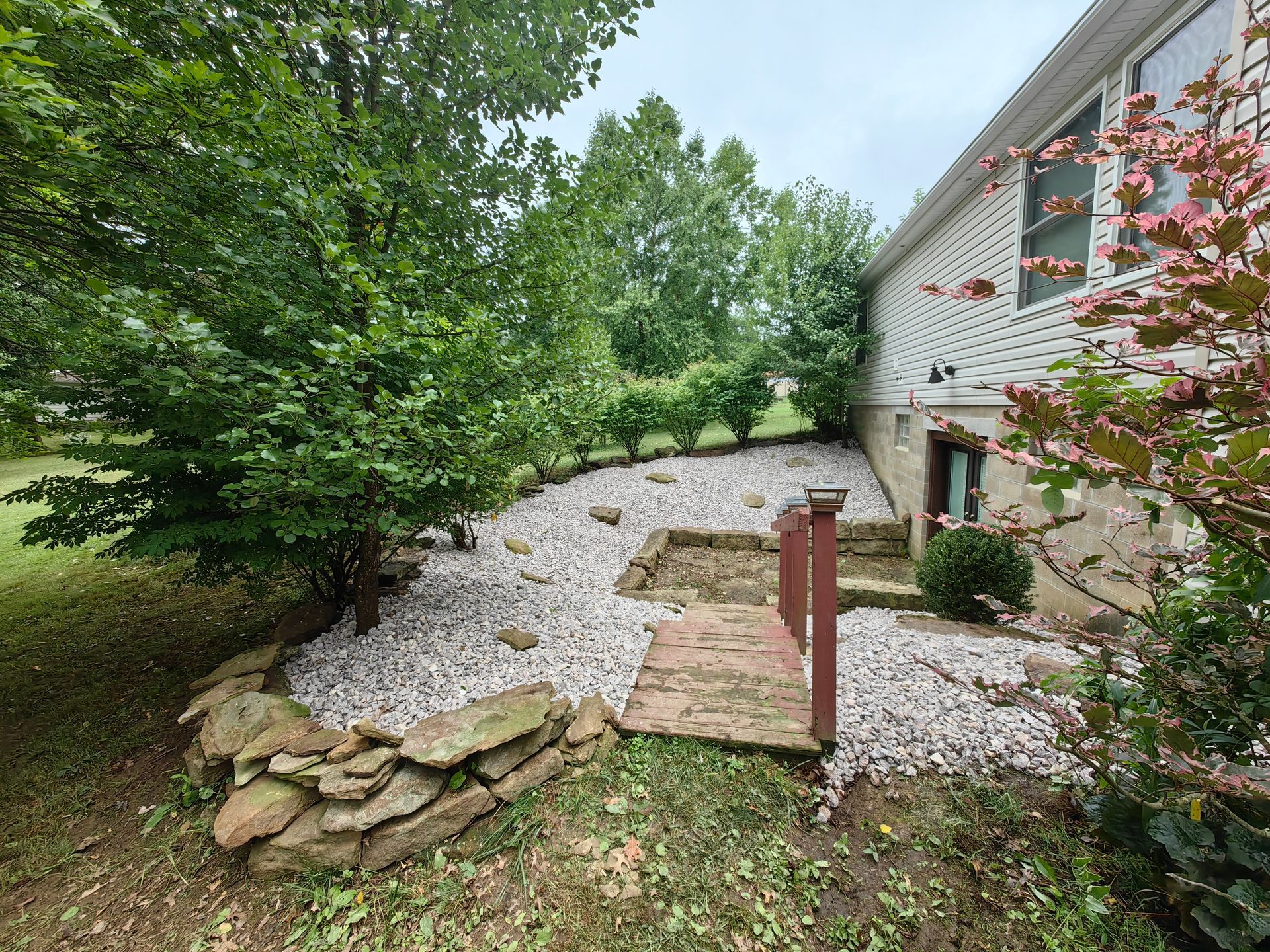 A backyard garden bed with white stones, stone steps, and a brick pathway next to a house.