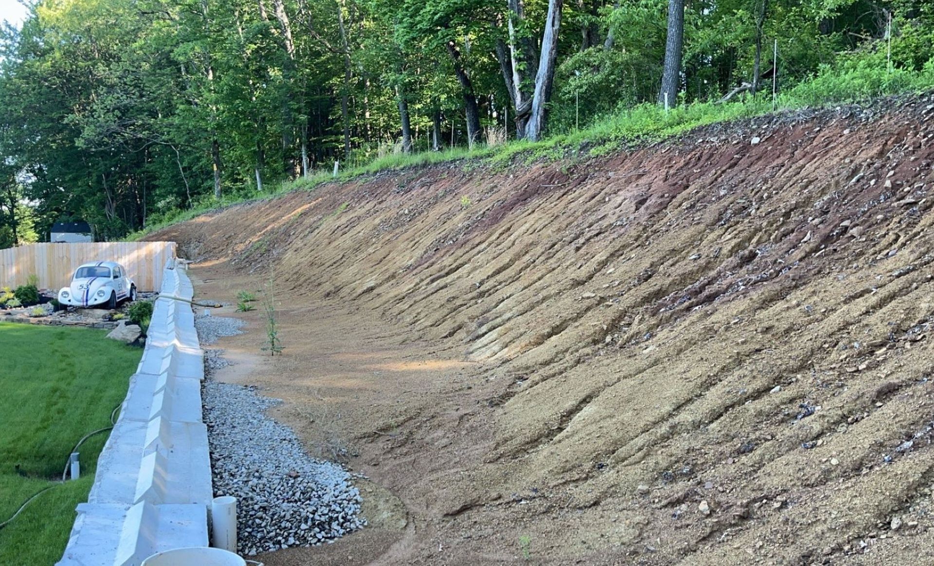 A sloped dirt hillside with a retaining wall. Green grass and trees are at the top and on the left.