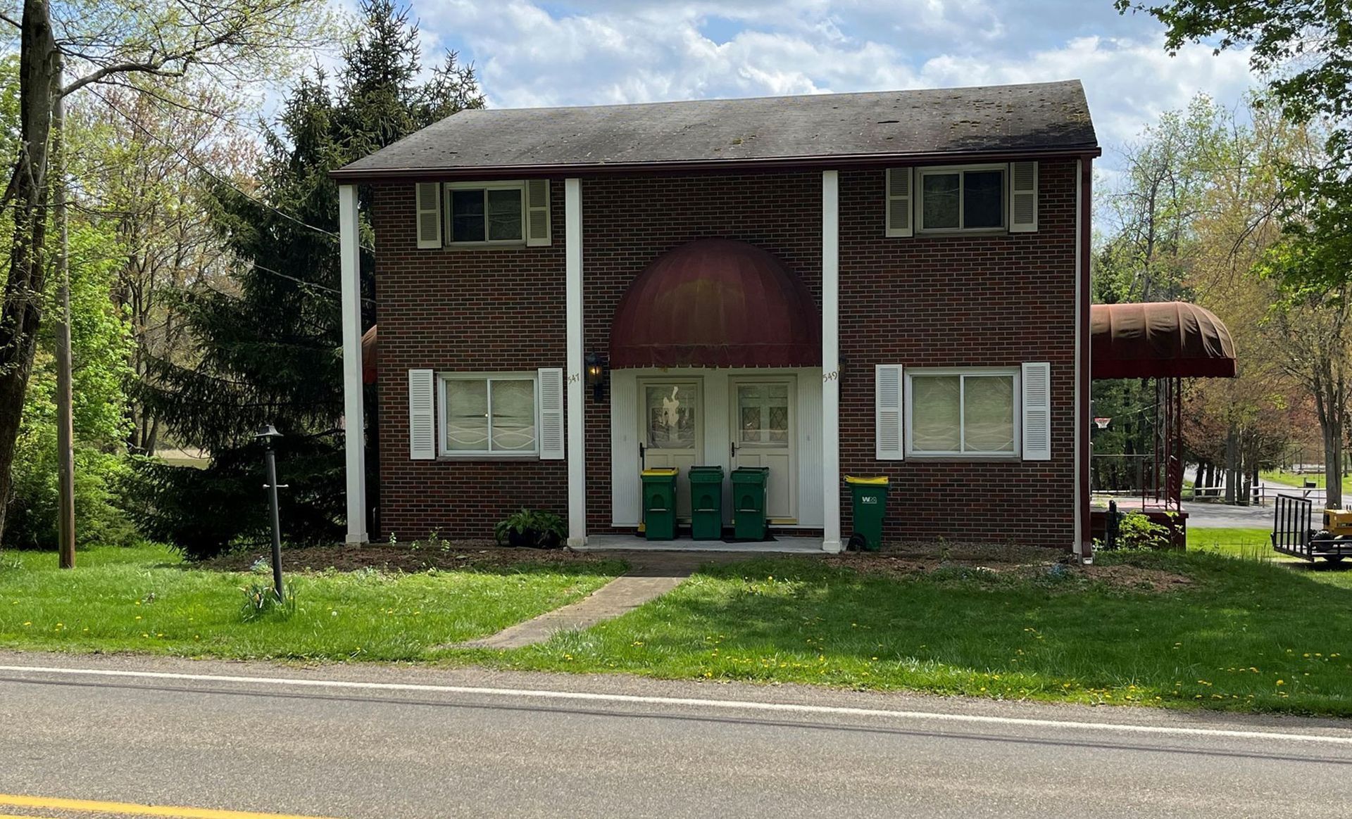 Two-story brown building with white trim, shutters, and a red awning, set on a grassy lot near a road.
