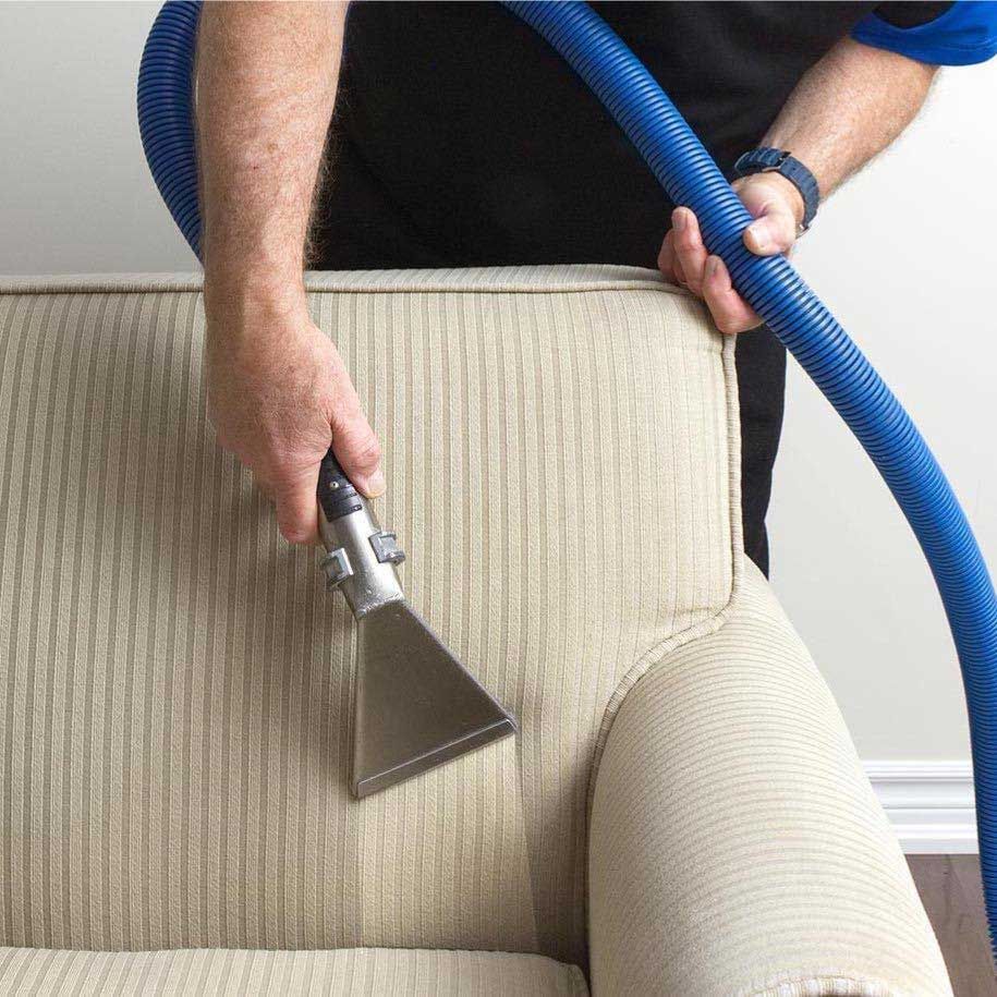 A person cleaning a beige corduroy sofa with a blue hose and cleaning head.