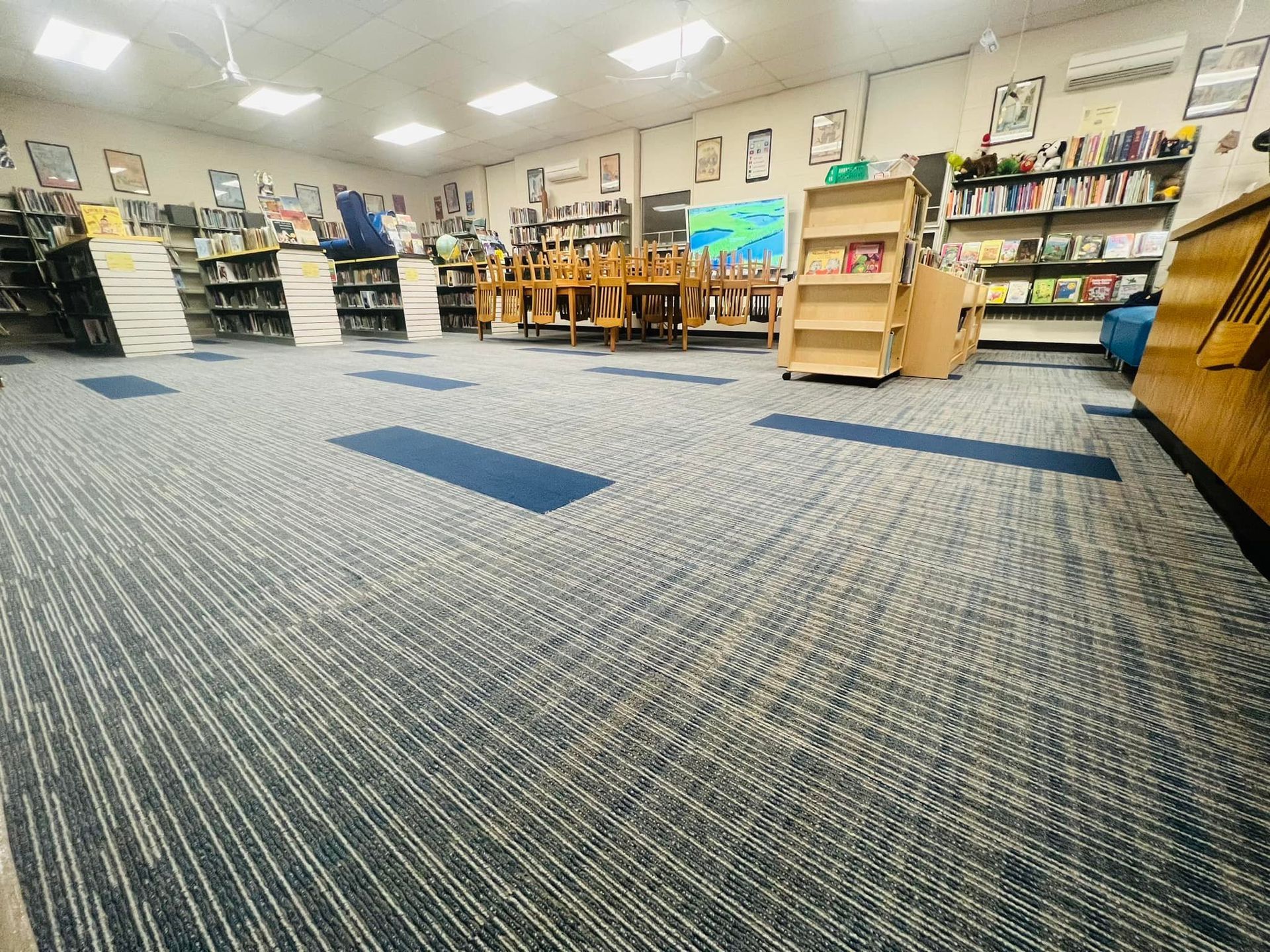 A library interior with blue and gray patterned carpet, books, tables, and shelves.
