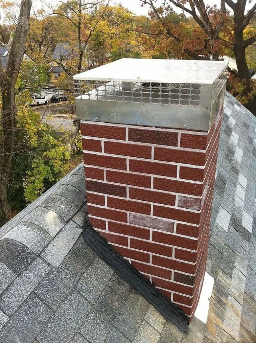 Brick chimney on a shingled roof with a metal cap, surrounded by autumn trees
