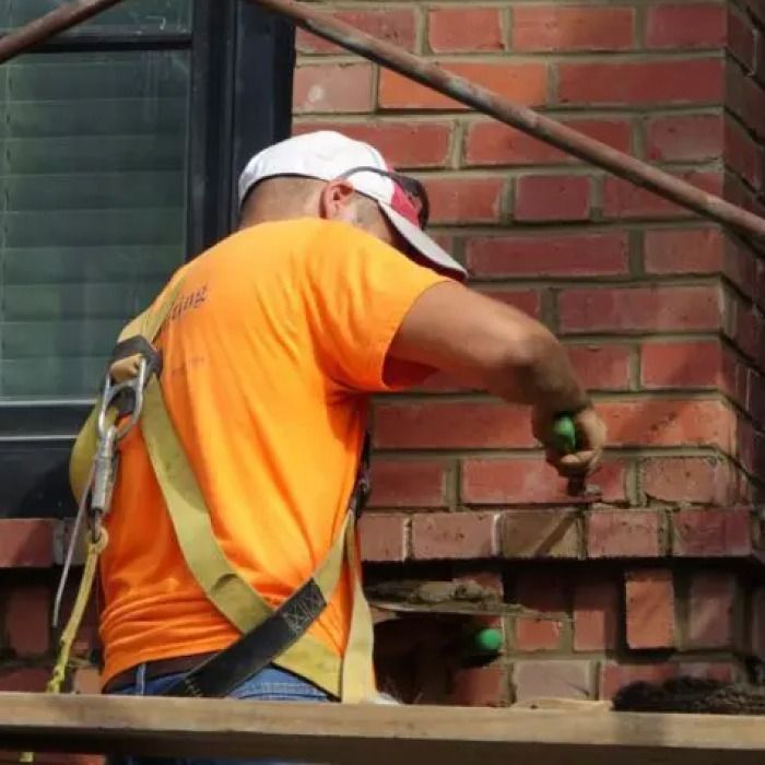 Construction worker in orange shirt repairs brickwork, wearing a safety harness.