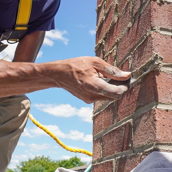 Person repairing brick chimney, using a tool to smooth mortar. Blue sky with clouds.