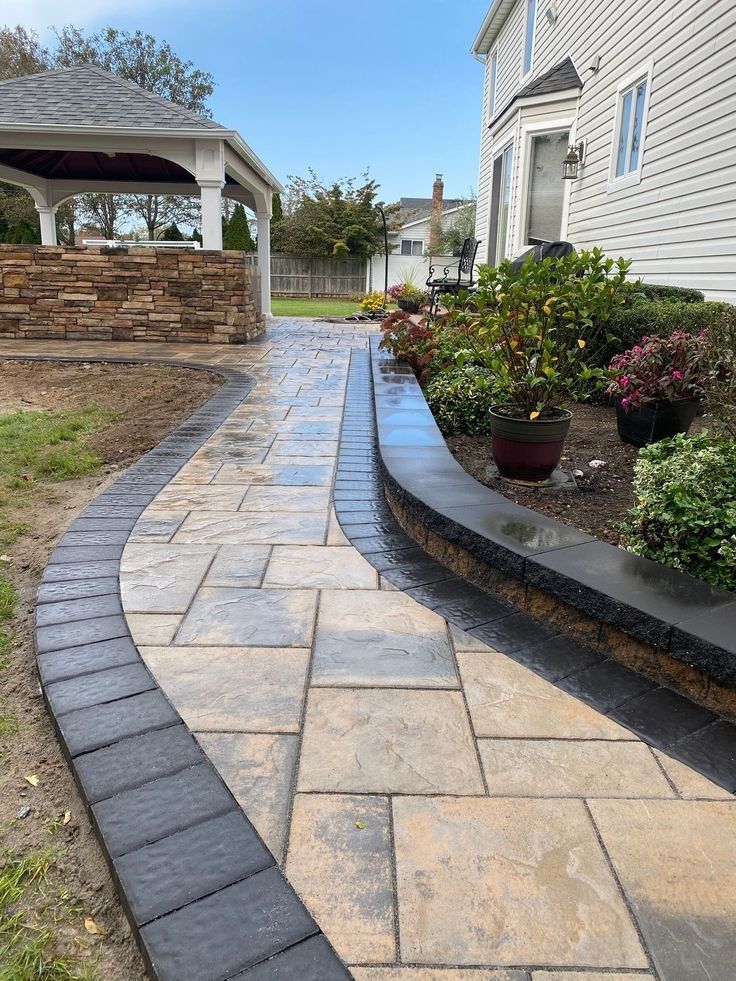 Paver pathway with retaining wall and garden leading to a gazebo beside a house.