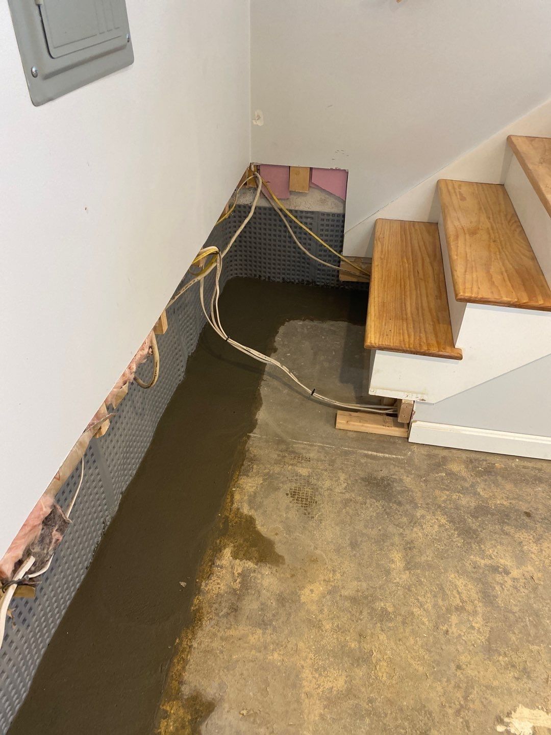 Basement under construction: concrete floor, wall with drainage membrane, wires, wooden stairs.