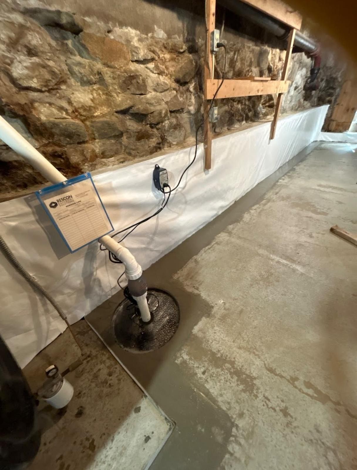 Basement with a sump pump system installed against a stone wall. White waterproof membrane covers the wall.