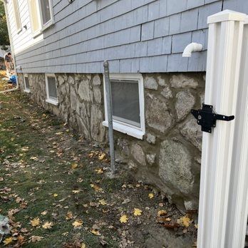 Stone foundation of a building with small windows. A white gutter is to the right. Leaves cover the ground.