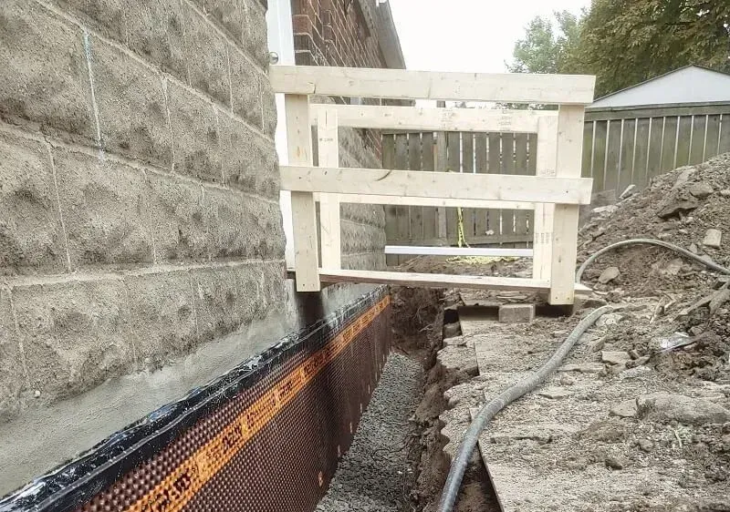Foundation repair: Wooden scaffolding above excavated trench beside a brick building and retaining wall.