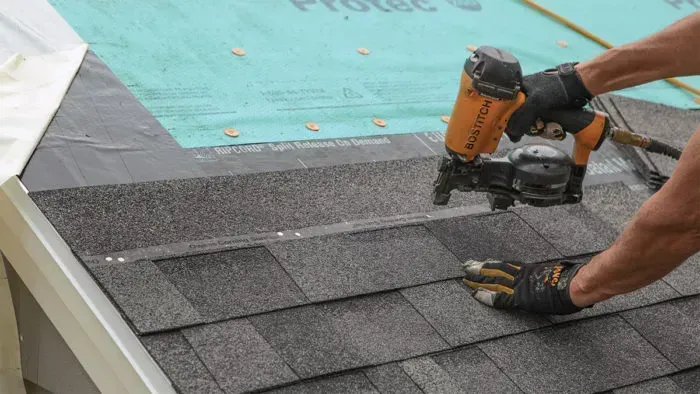 A roofer uses a nail gun to install asphalt shingles on a roof.