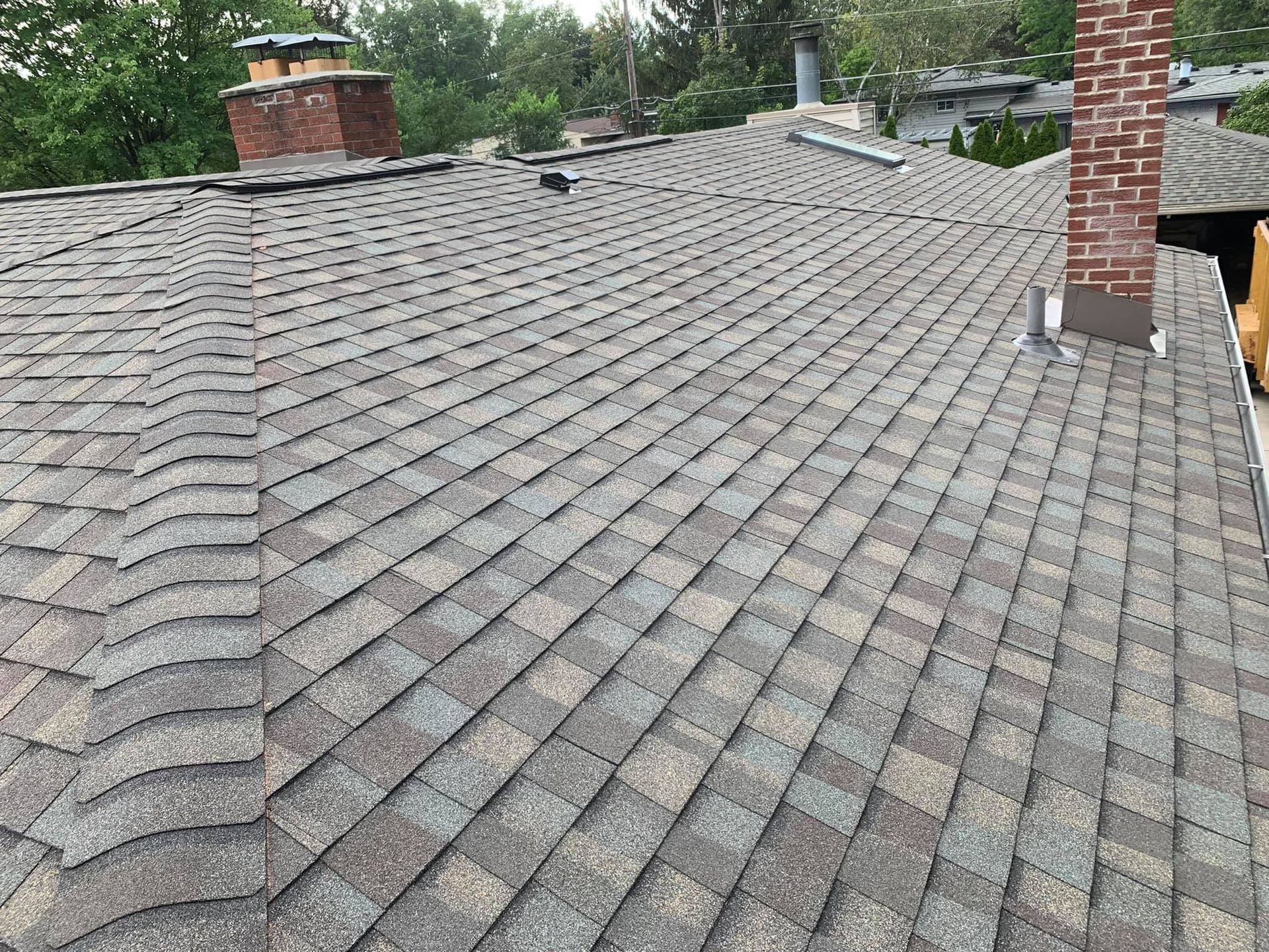 Gray asphalt shingle roof with two brick chimneys against a backdrop of green trees and a partly cloudy sky.