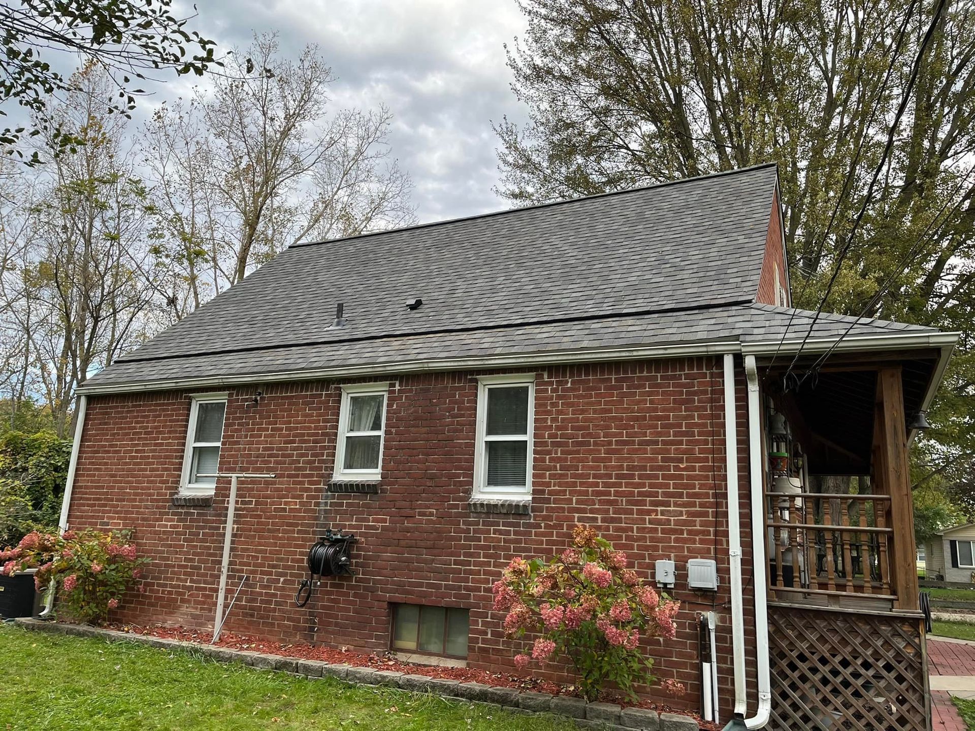 Brick house with gray roof and porch, surrounded by greenery under a cloudy sky.