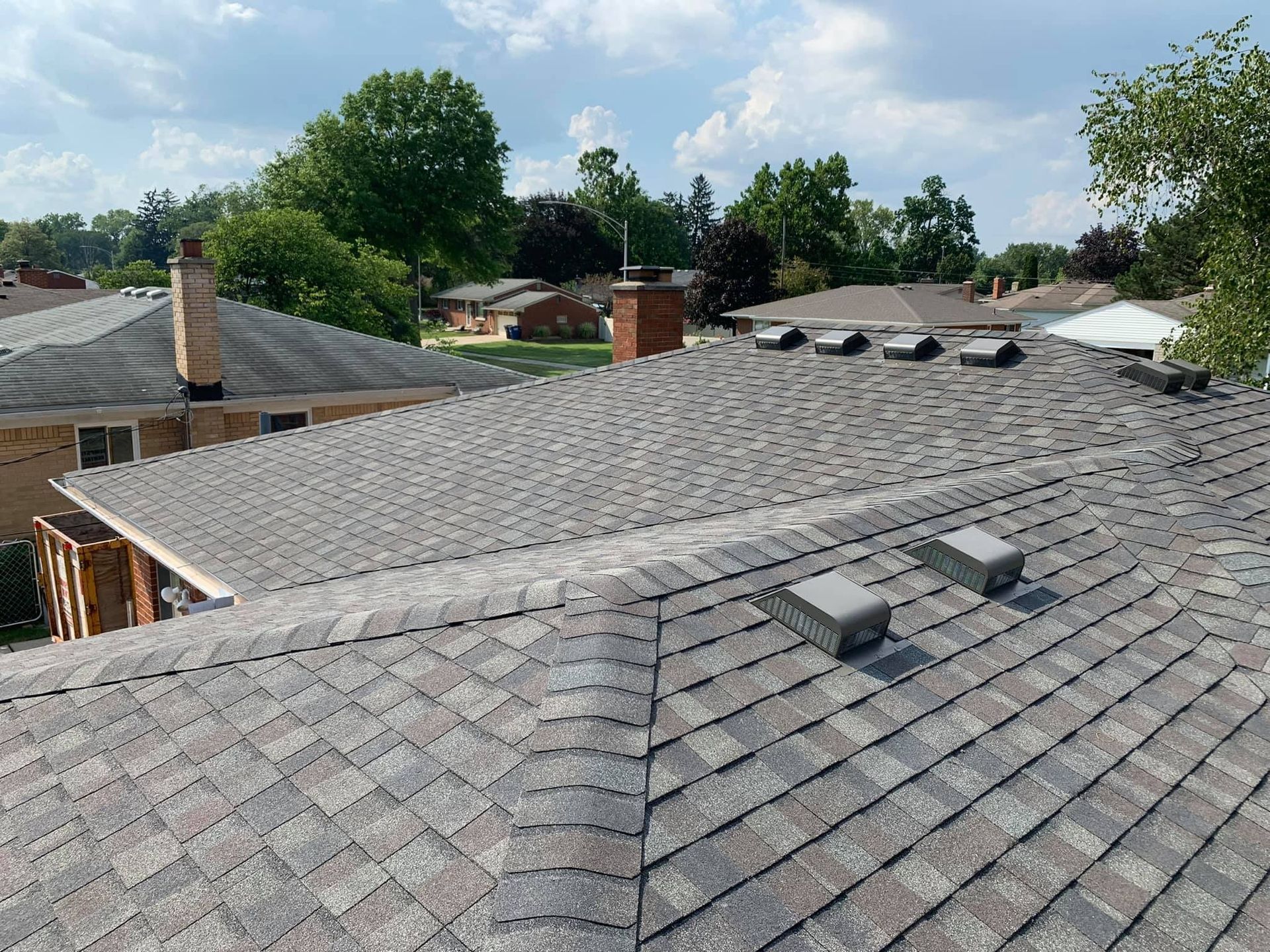 View of a house roof covered in gray and brown shingles. Chimney, vents, and surrounding houses in background.