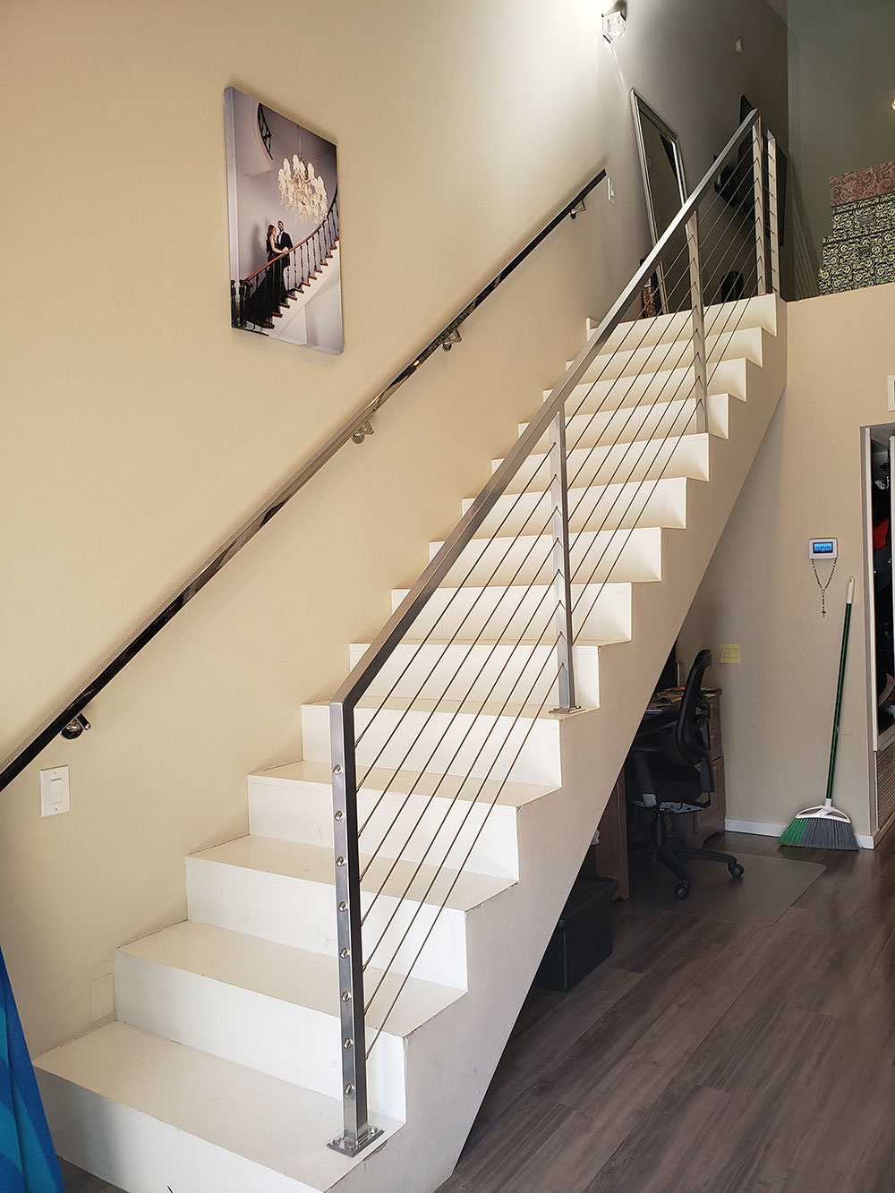 Staircase with white steps, stainless steel railing with cable infill, black handrail on beige wall.