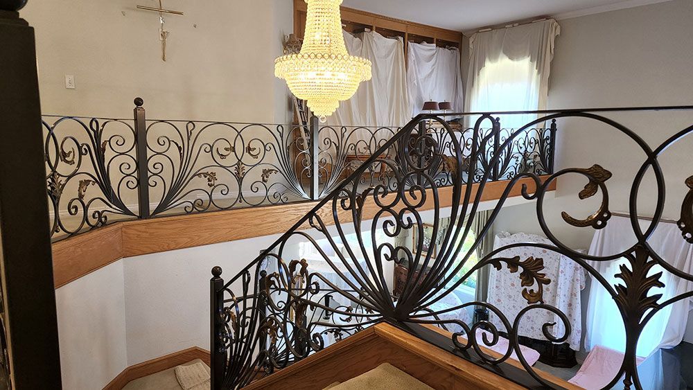 Ornate wrought-iron staircase with a chandelier. Beige walls, wood accents, and light from a window.