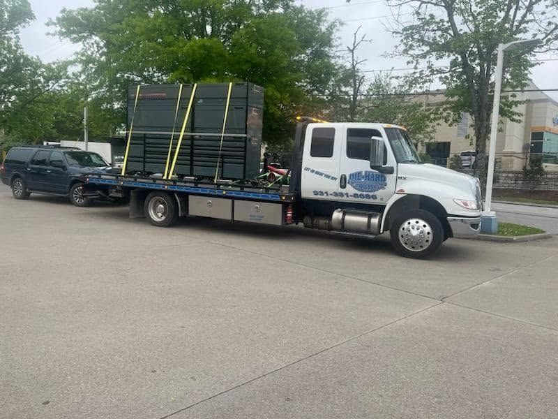 White flatbed tow truck carrying stacked black crates on a street, with a dark SUV behind it