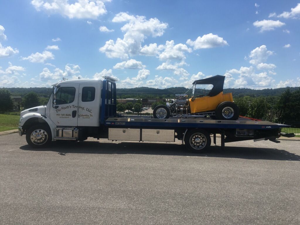 Tow truck carrying a yellow skid steer on a flatbed, parked under a bright blue sky.