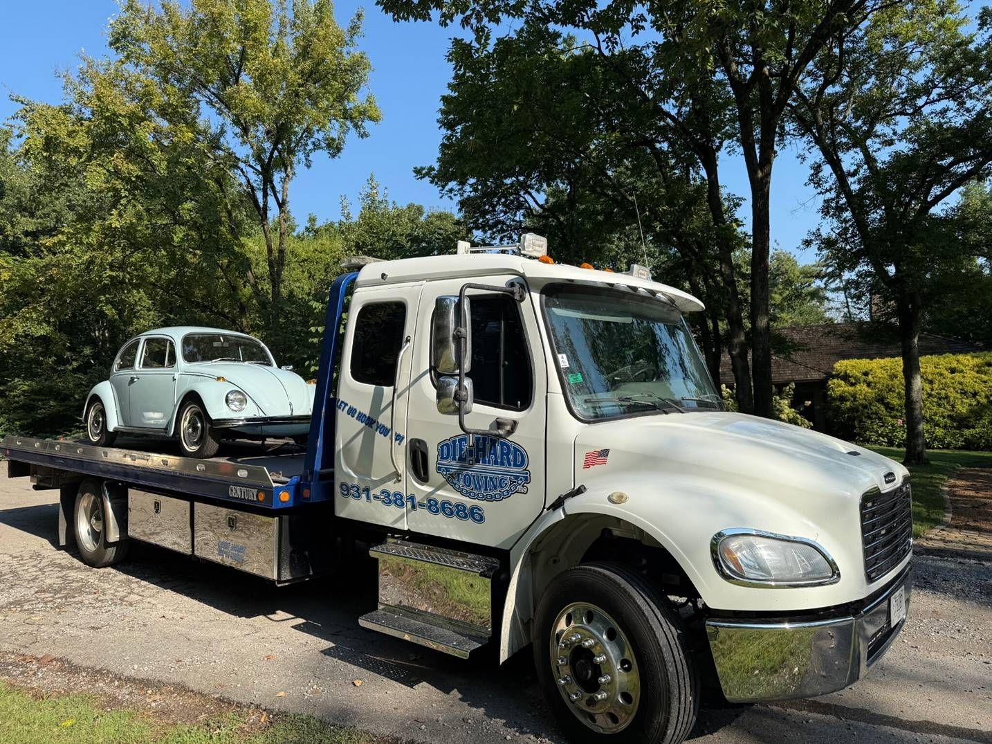 White flatbed tow truck carrying a light blue classic car on a roadside with trees