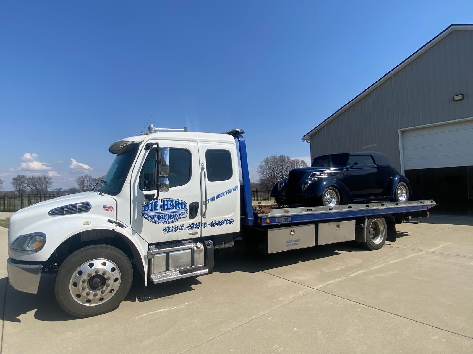 White tow truck hauling a black car beside a warehouse on a sunny day