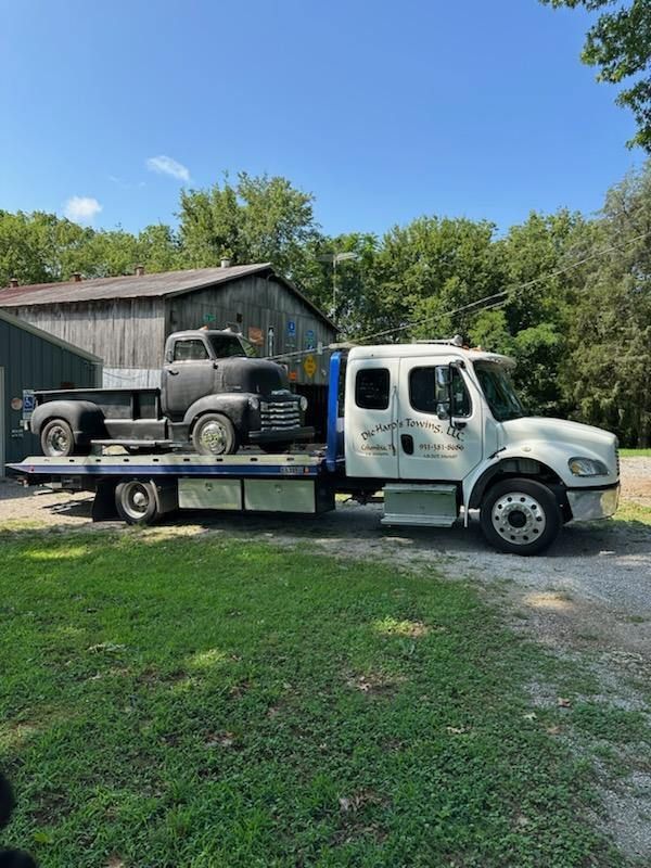 White flatbed tow truck carrying a vintage Jeep beside a rustic barn on a sunny grassy farm.