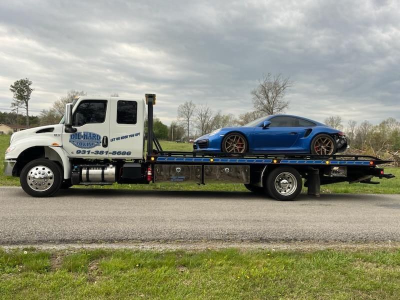 Blue sports car on a tow truck driving along a roadside under a cloudy sky
