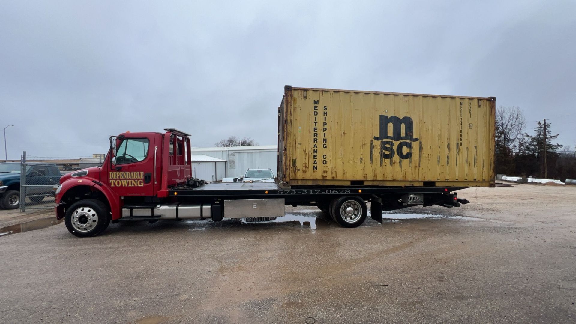 Red semi-truck hauling a yellow shipping container in a gravel lot under a cloudy sky