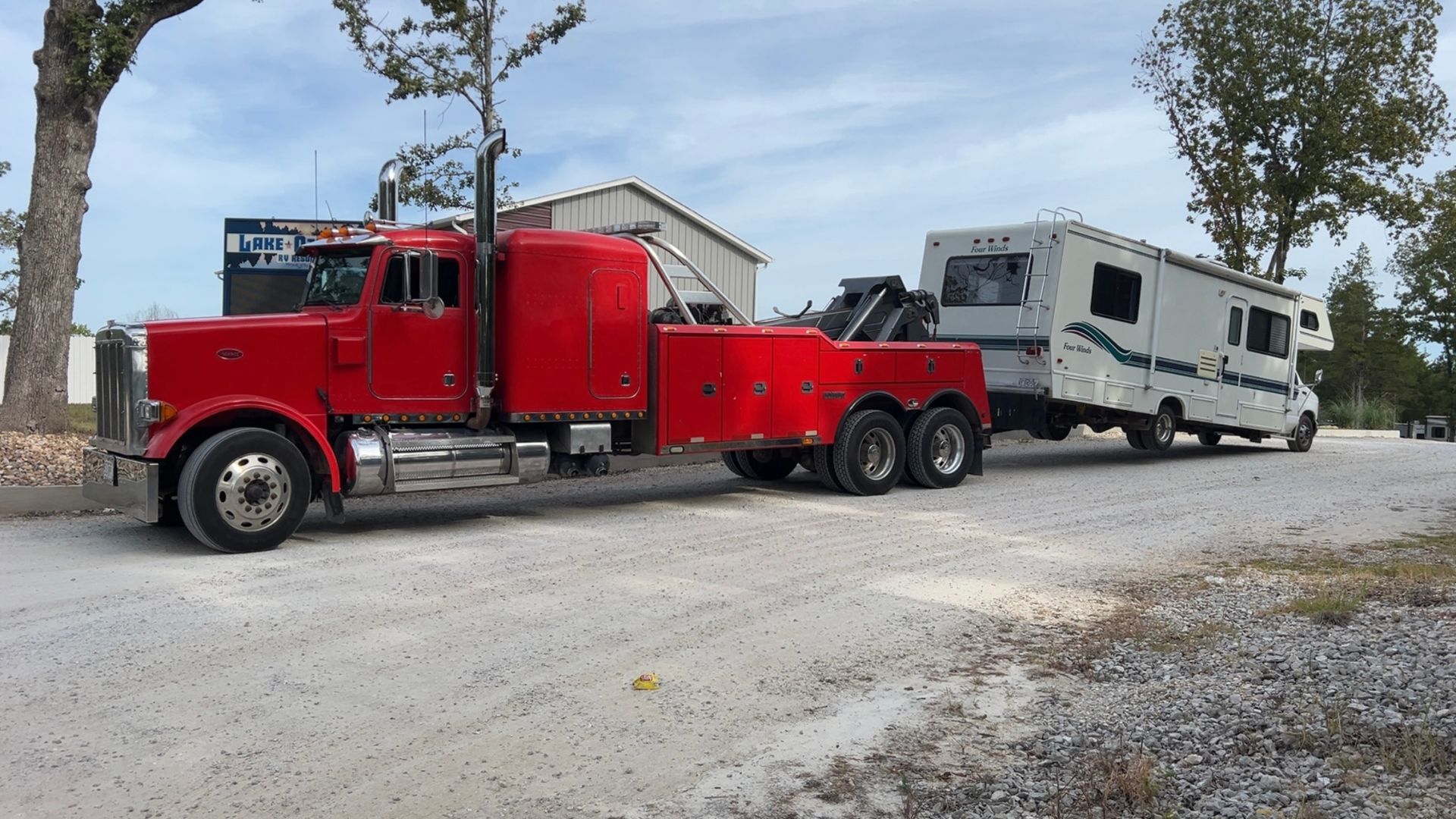 Red tow truck hauling a white RV on a gravel road under a cloudy sky