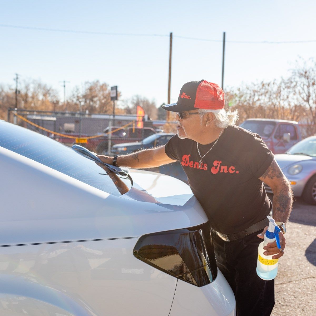 A man wearing a black shirt that says burts inc. is cleaning the windshield of a white car