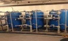 Blue industrial water filtration tanks in a row with pipes and gauges, indoors.