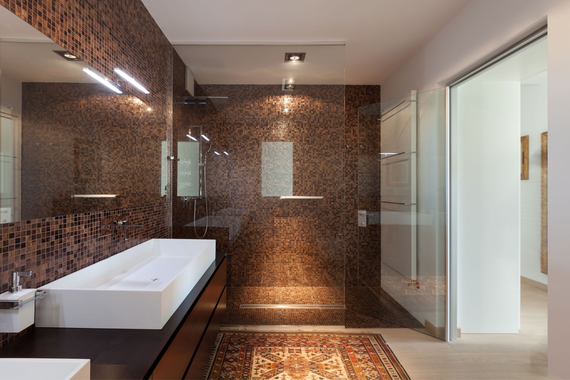 Modern bathroom with brown mosaic tile, white sink, glass shower, and patterned rug.
