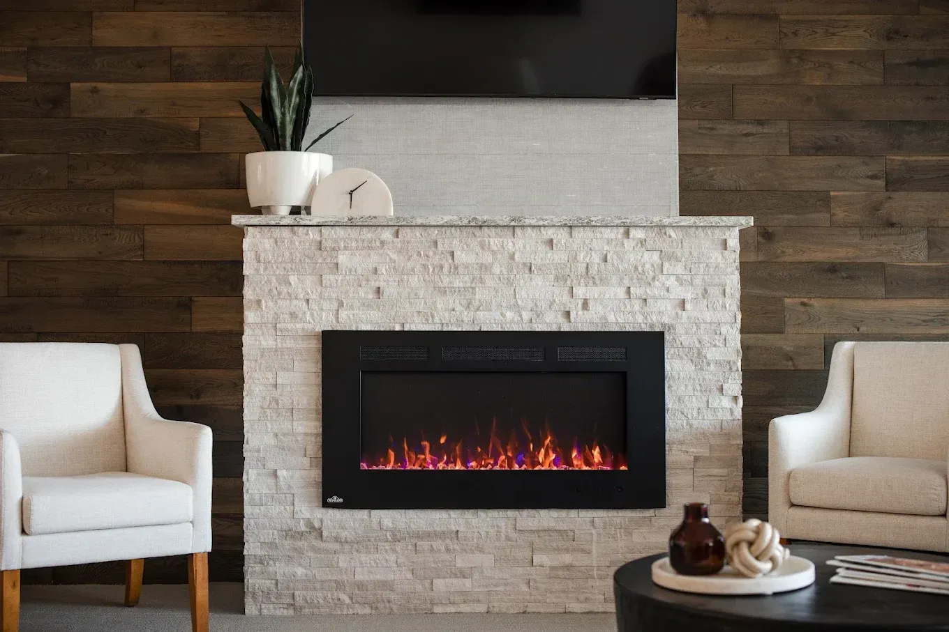 Modern fireplace with a black frame and faux flame, flanked by white chairs and dark wood wall.