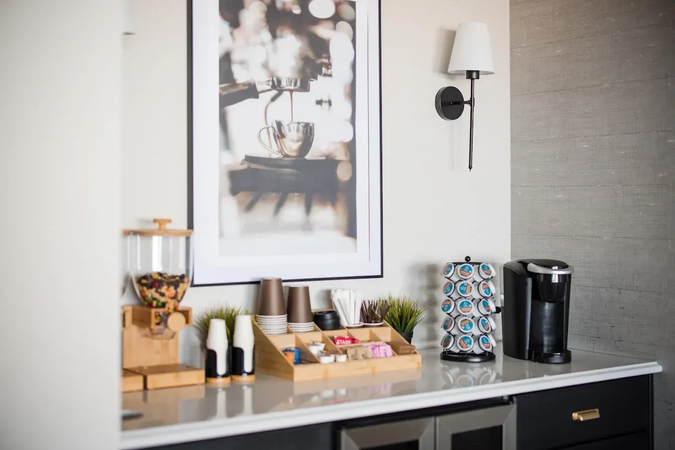 Coffee station with dispenser, Keurig, cups, and art print on a counter.