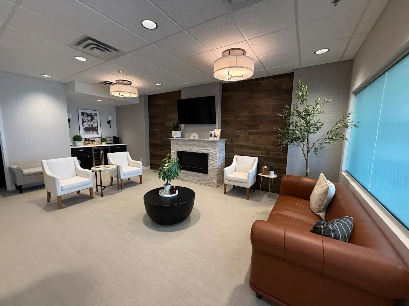 Lounge area with seating, fireplace, and bar. Neutral colors with wood accent wall and windows.