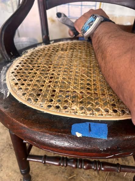 Person repairing a woven cane chair seat with a tool.