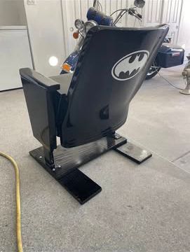 Batman-themed barber chair; black and silver; bat symbol on backrest; concrete floor; motorcycle in background.