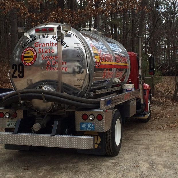 A red septic tank truck parked on a gravel road, with trees in the background. The tank is silver and has company logos.