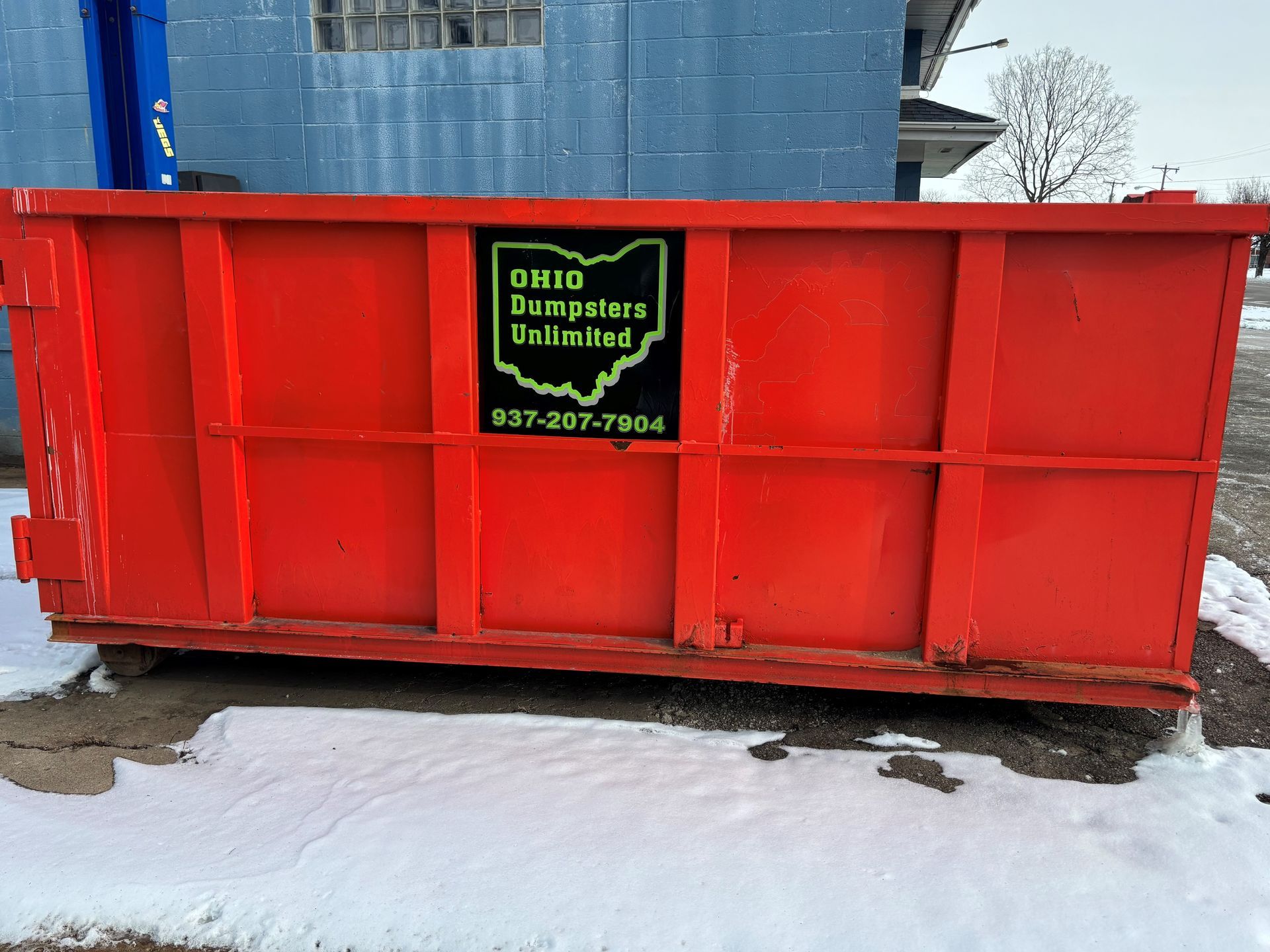 Red Ohio Dumpsters Unlimited dumpster in front of a blue building, with patches of snow on the ground.