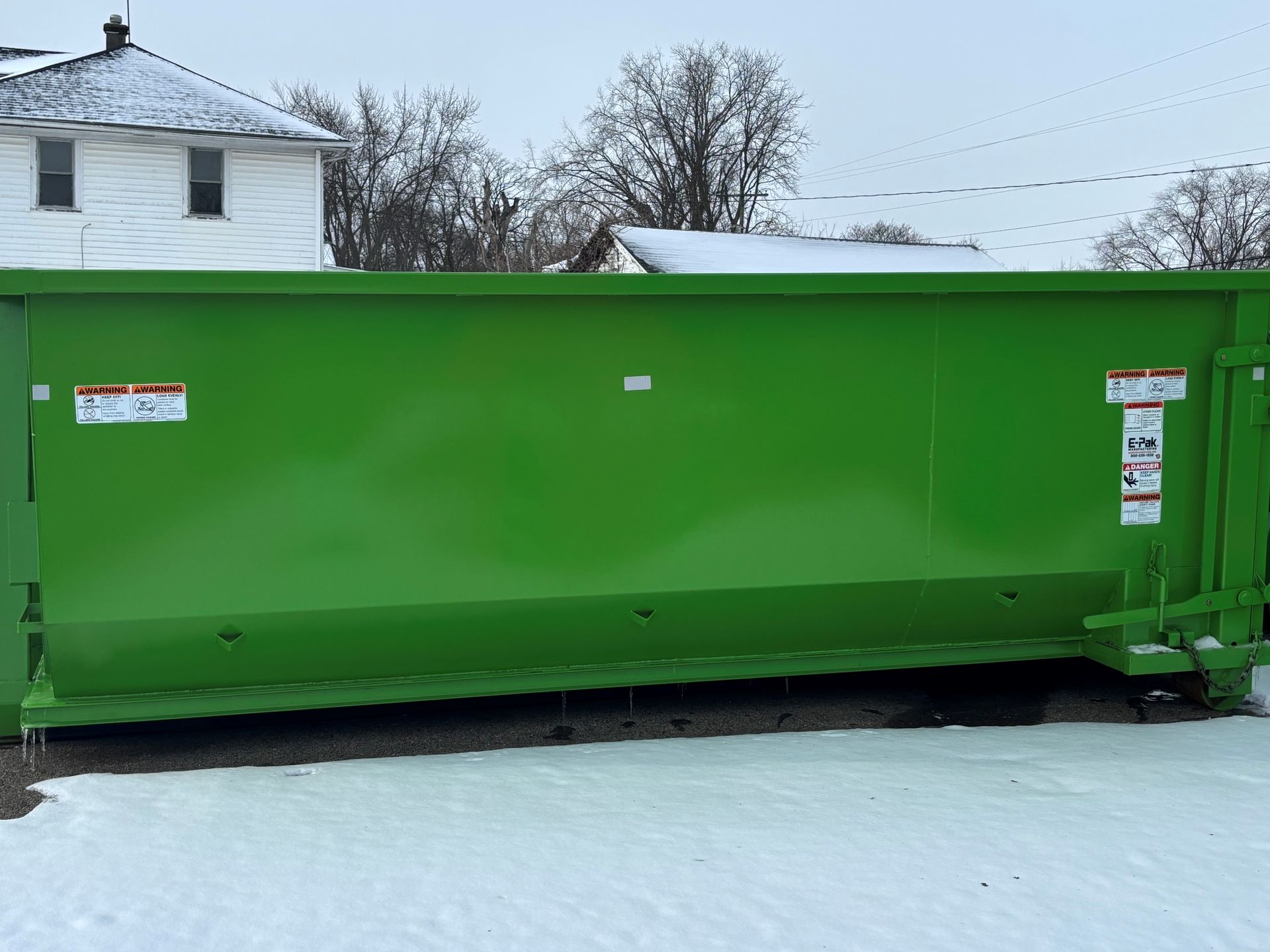 Green dumpster in a snowy setting with a house and trees visible in the background.