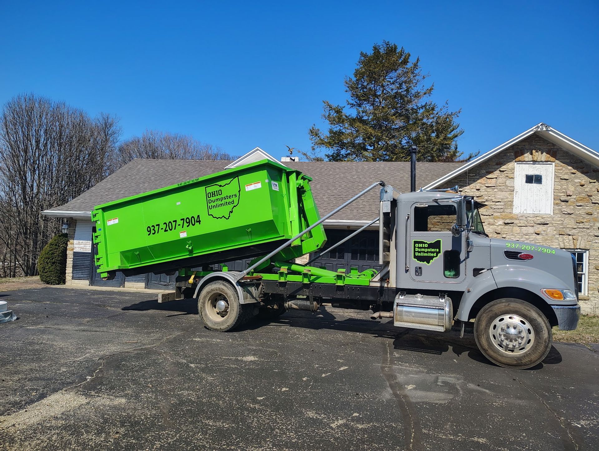 Green dumpster being lifted by a gray truck in front of a stone house on a sunny day.