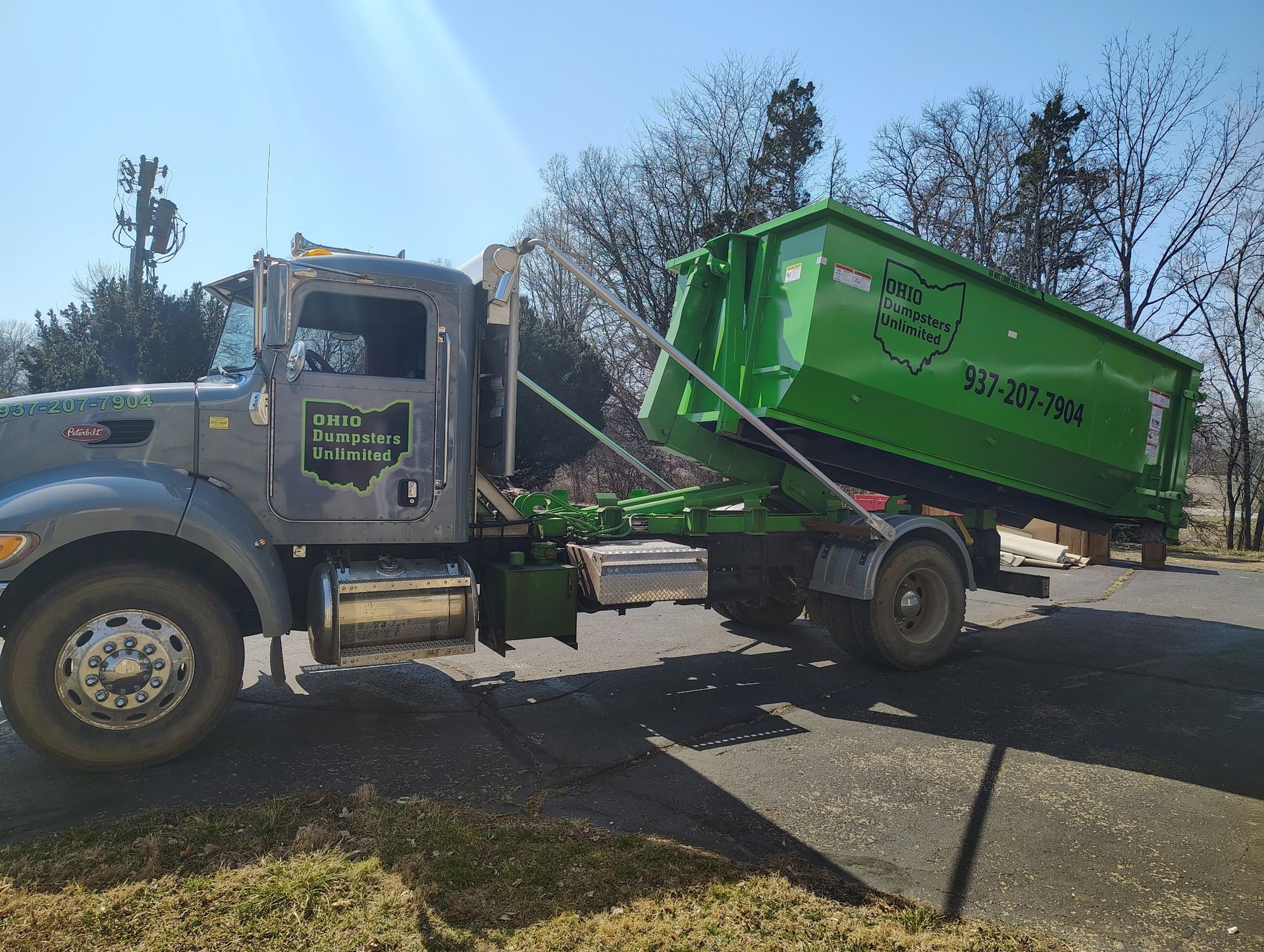 Gray truck with green dumpster in an outdoor setting, angled up to dump.