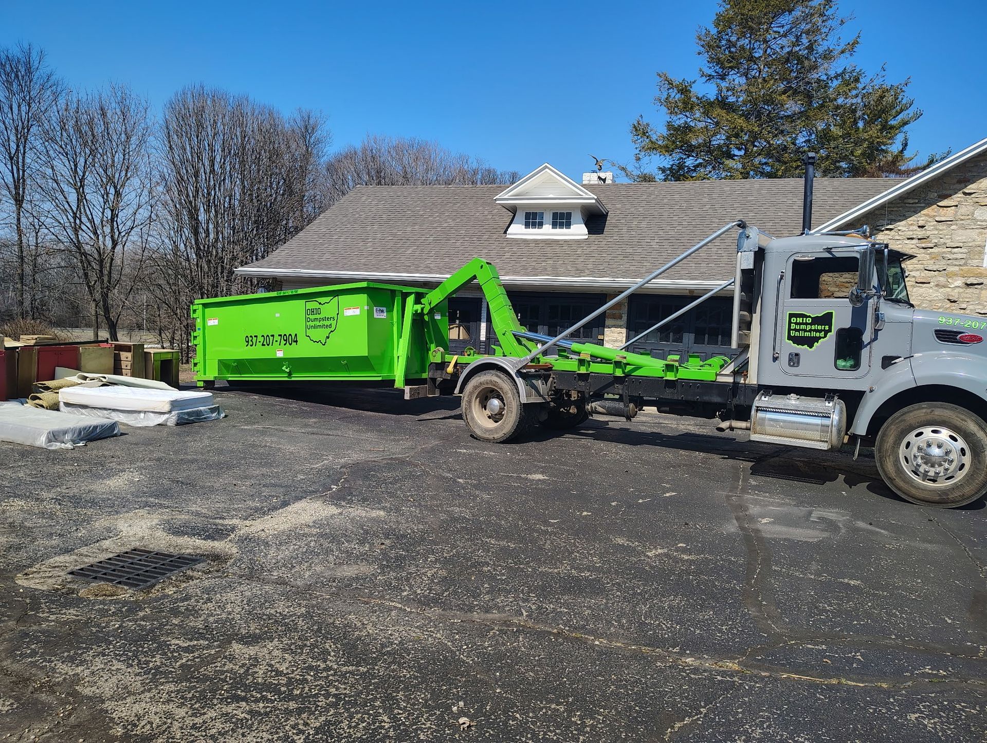 Green dumpster being loaded onto a gray truck in front of a house on a sunny day.
