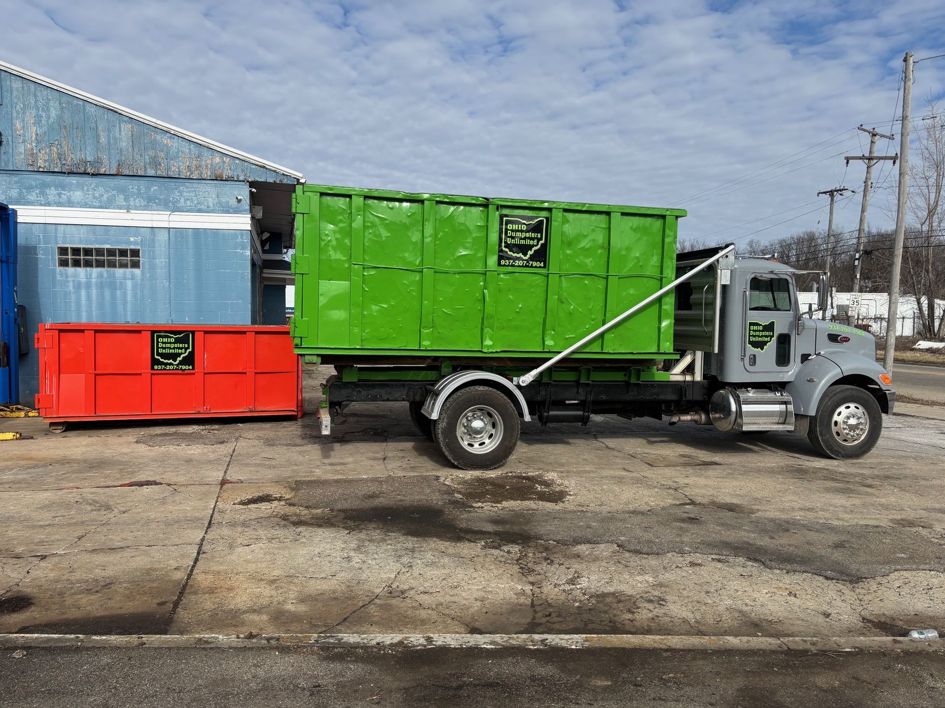 Green dumpster on a gray truck with a red dumpster nearby, set on concrete, next to a blue building.