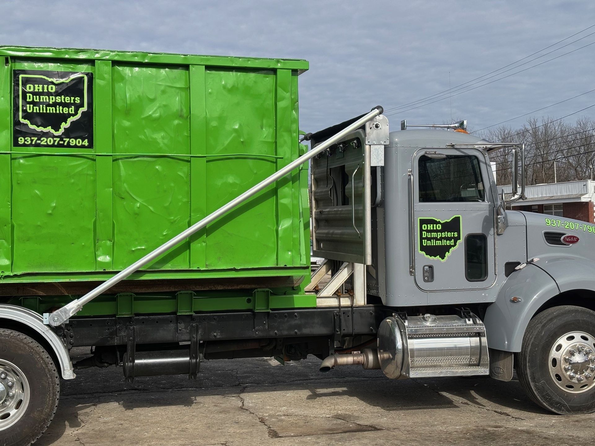 Green dumpster loaded on a gray Ohio Permian Truck, under a cloudy sky.