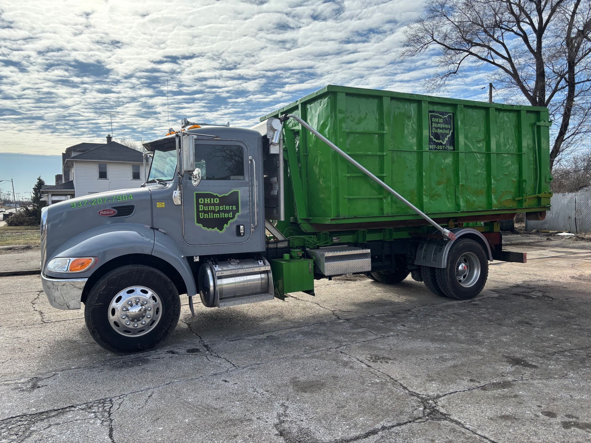 Gray dump truck with a green dumpster on a paved lot under a cloudy sky.