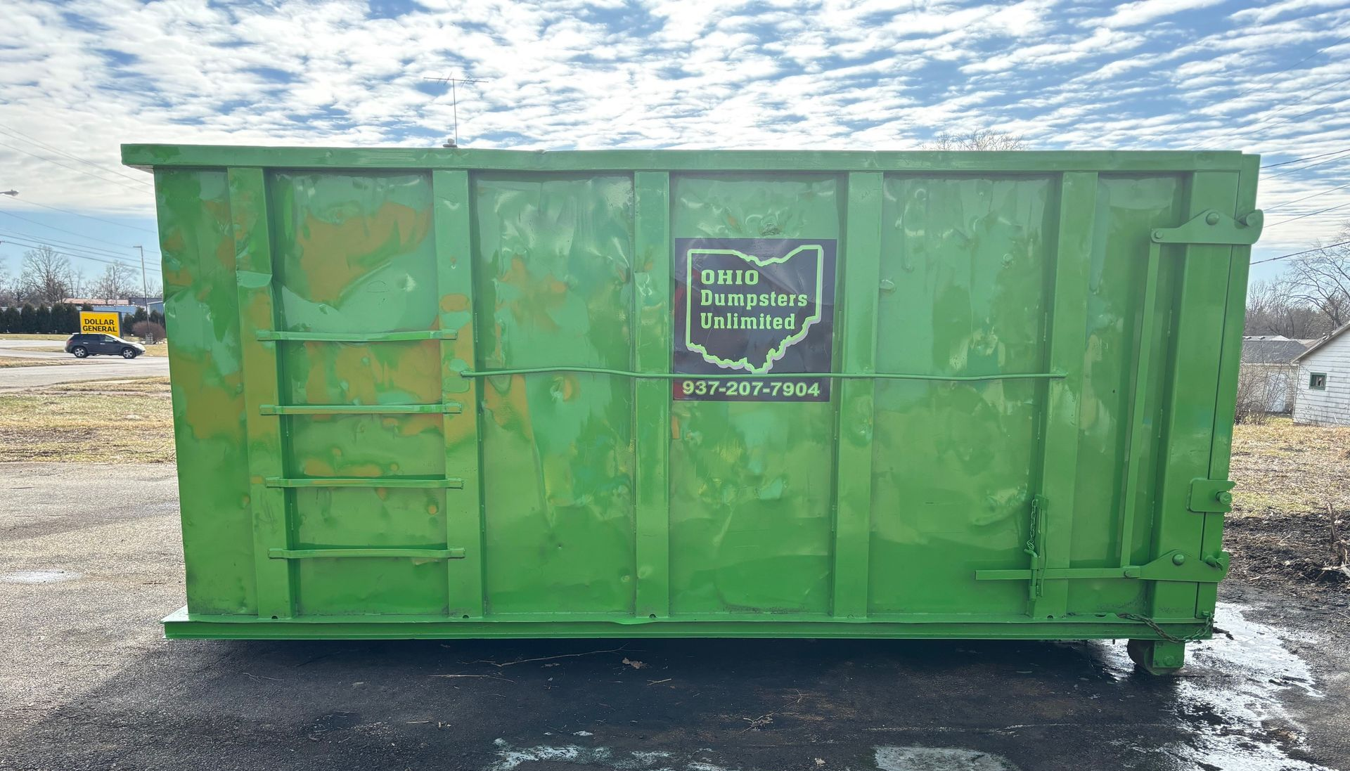 Green dumpster on a truck in front of a house on a sunny day.