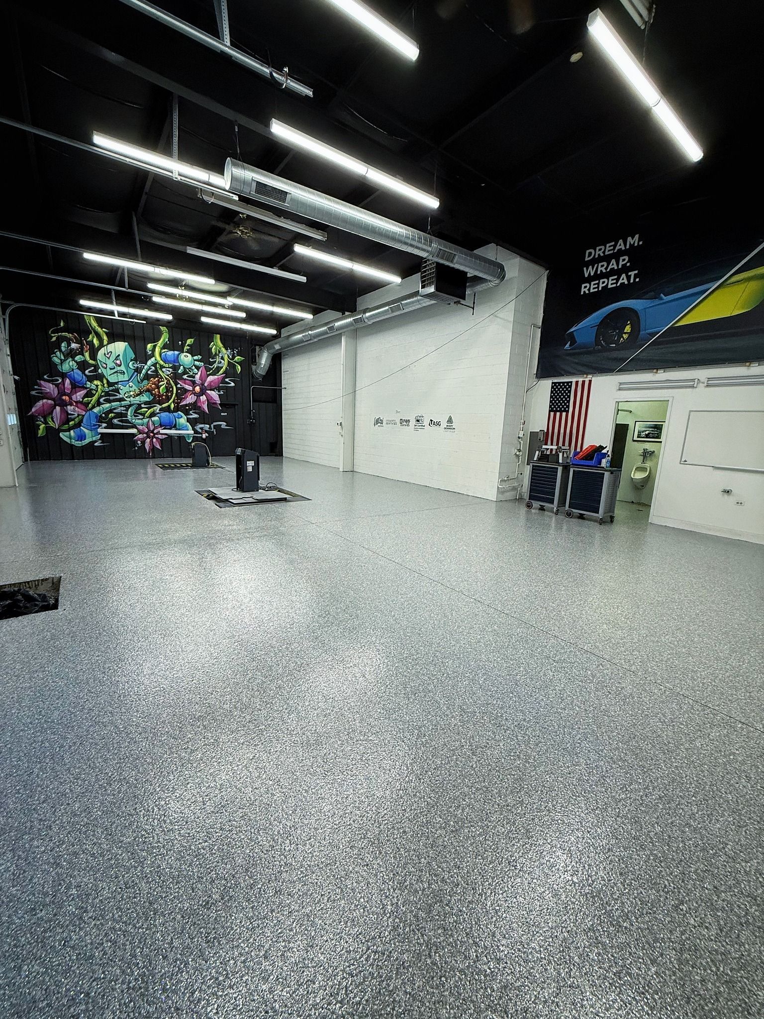 Spacious room with a speckled gray floor, black ceiling, white walls, and a colorful mural. Bright lights illuminate the space.