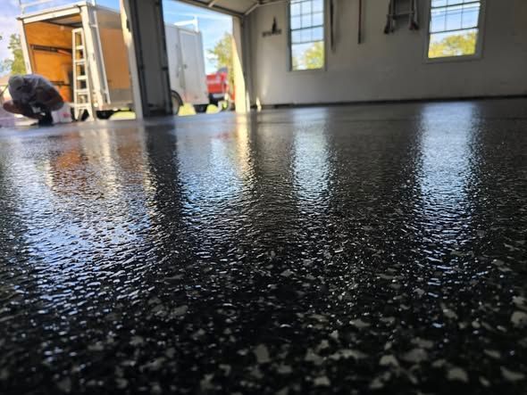 Shiny, speckled epoxy floor inside a garage, open to the outdoors. A person is working in the background.