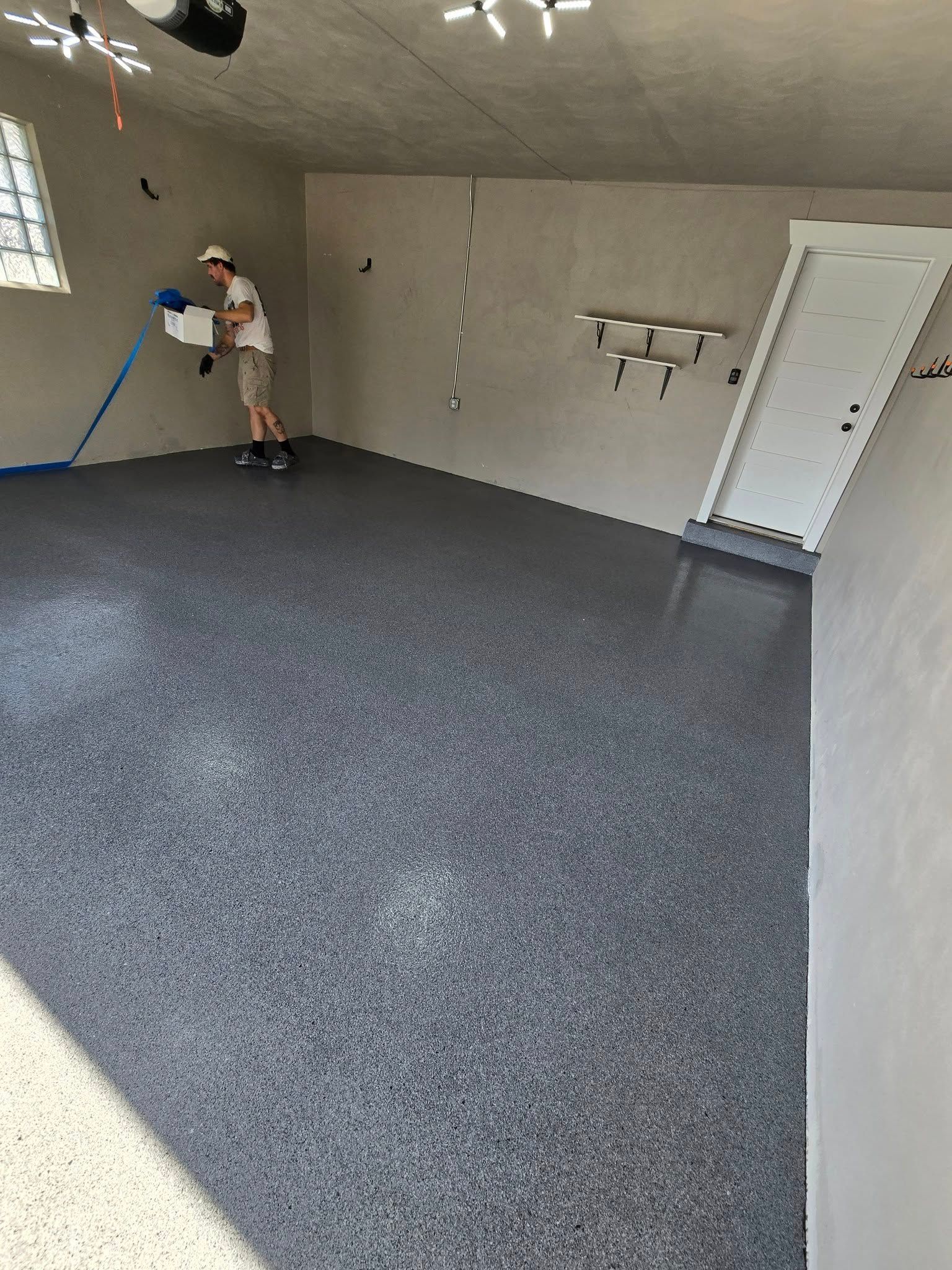 Person applying epoxy flooring in a garage. Floor is speckled gray, walls are light gray.