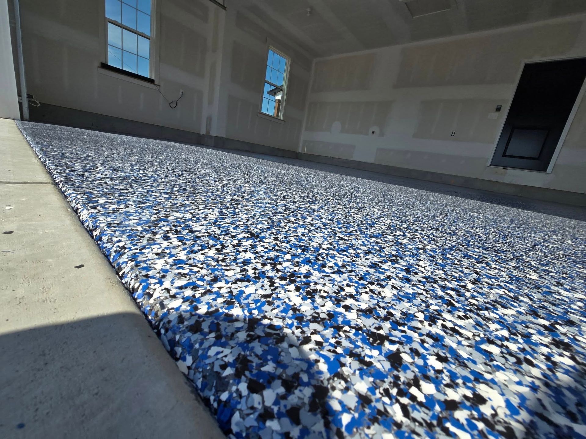 Garage floor with blue, white, and black flecks. Concrete ramp leads to interior with drywall and windows.