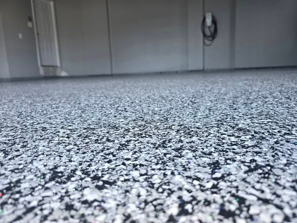 Close-up of a garage floor with a speckled epoxy coating, gray and white chips. A charger is mounted on the wall.